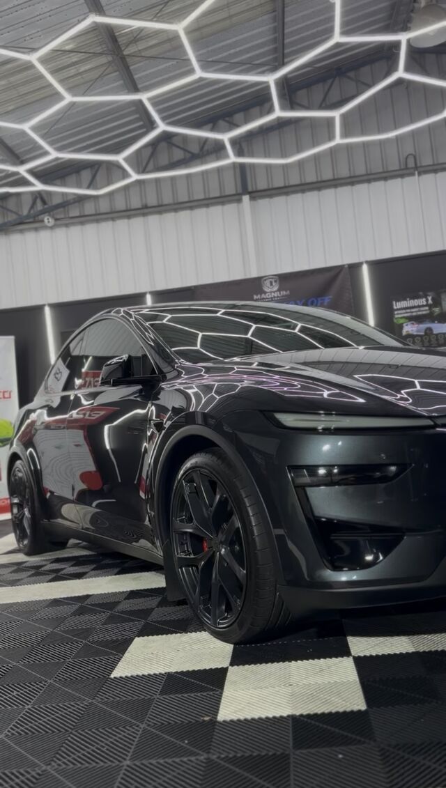 Dark grey performance coupe under the hexagon inspection lighting, its ceramic-coated paintwork mirroring the lights